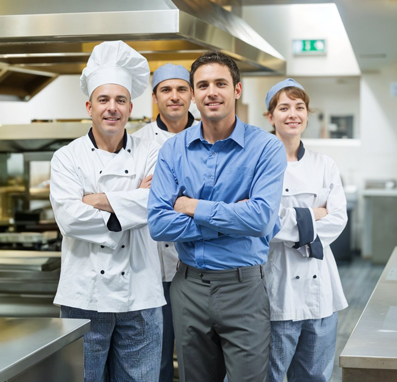 Vier Personen in einer Restaurantküche: drei in weißen Kochjacken und Kochmützen sowie eine Person im blauen Hemd stehen mit verschränkten Armen und lächeln.