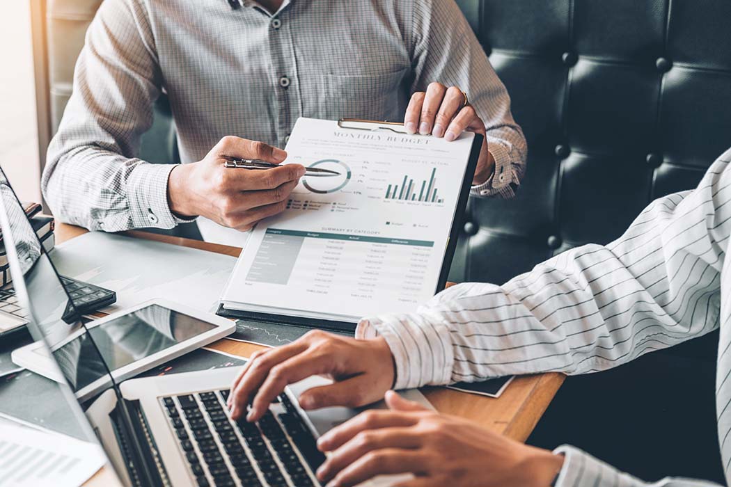 Zwei Personen in Hemden an einem Tisch: eine zeigt mit einem Stift auf ein Klemmbrett mit Kreis- und Balkendiagrammen und Tabellen, die andere tippt auf einem Laptop, neben Tablets und Unterlagen liegen Geräte und Papiere.