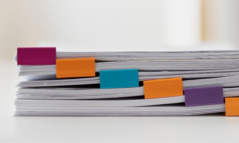 Mehrere gestapelte Papierbündel sind an den Kanten mit bunten rechteckigen Clips in Rosa, Orange, Türkis und Lila befestigt vor hellem, unscharfem Hintergrund.