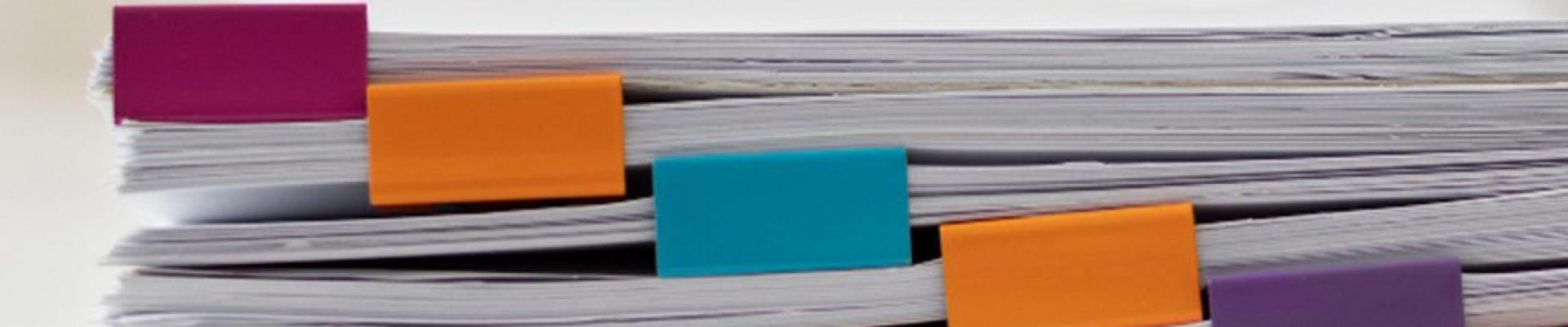 Mehrere gestapelte Papierbündel sind an den Kanten mit bunten rechteckigen Clips in Rosa, Orange, Türkis und Lila befestigt vor hellem, unscharfem Hintergrund.