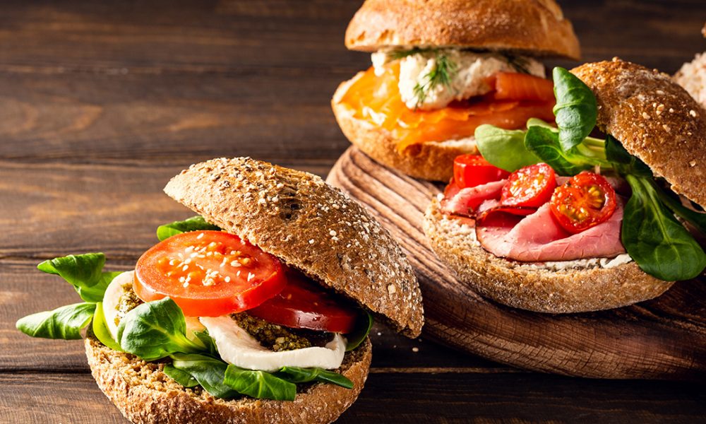 Drei rustikale Vollkornbrötchen auf einem Holzbrett mit verschiedenen Belägen: vorn Mozzarella, Pesto, Tomatenscheiben und grünes Blattgemüse, dahinter Räucherlachs mit Dillcreme und Roastbeef mit Kirschtomaten und Salat.