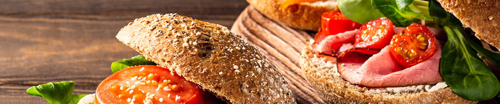 Drei rustikale Vollkornbrötchen auf einem Holzbrett mit verschiedenen Belägen: vorn Mozzarella, Pesto, Tomatenscheiben und grünes Blattgemüse, dahinter Räucherlachs mit Dillcreme und Roastbeef mit Kirschtomaten und Salat.