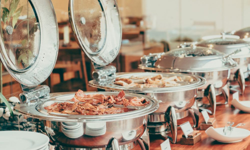 Mehrere glänzende Edelstahl-Warmhalteschalen mit geöffneten, beschlagenen Deckeln auf einem Holztisch, gefüllt mit Frühstücksgerichten wie gebratenem Speck und Würstchen, daneben gestapelte weiße Teller und Servierbesteck.