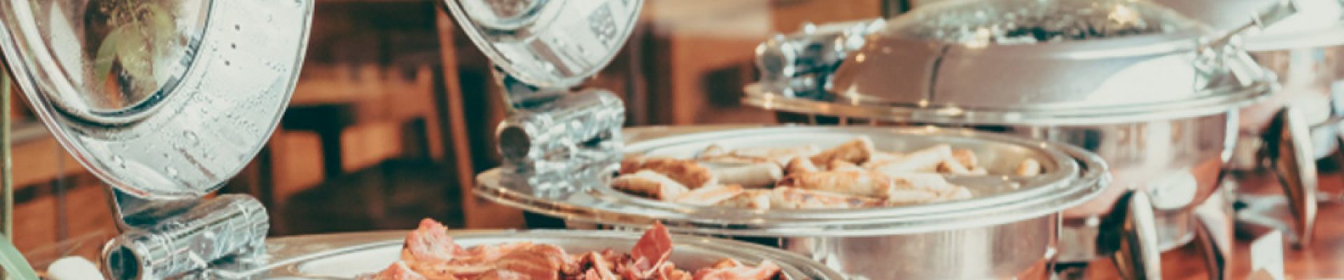 Mehrere glänzende Edelstahl-Warmhalteschalen mit geöffneten, beschlagenen Deckeln auf einem Holztisch, gefüllt mit Frühstücksgerichten wie gebratenem Speck und Würstchen, daneben gestapelte weiße Teller und Servierbesteck.