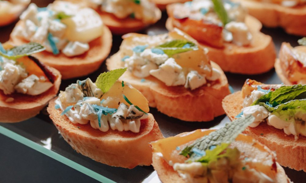 Mehrere Crostini auf einem Glas-Serviertablett: geröstete Baguettescheiben belegt mit weichem Käse und Blauschimmelkrümeln, karamellisierter Birne und frischen Minzblättern.