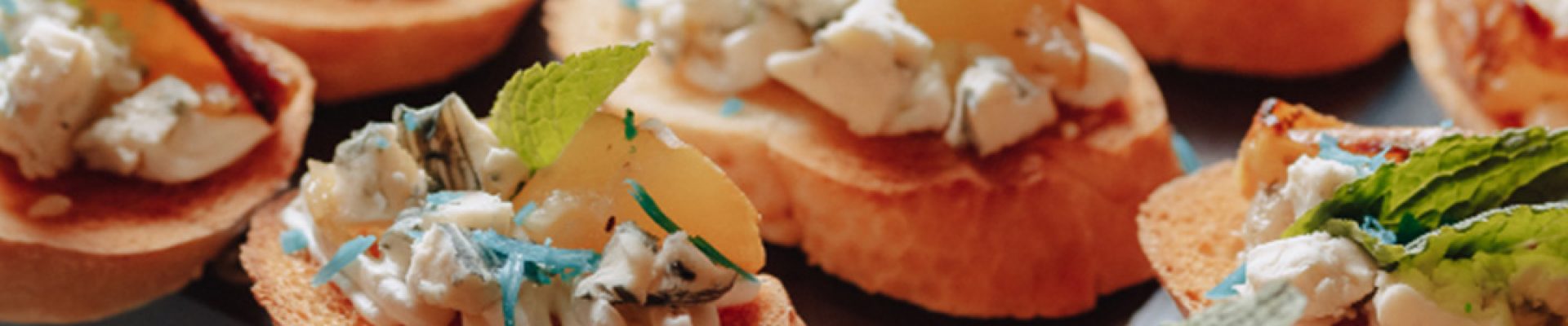 Mehrere Crostini auf einem Glas-Serviertablett: geröstete Baguettescheiben belegt mit weichem Käse und Blauschimmelkrümeln, karamellisierter Birne und frischen Minzblättern.