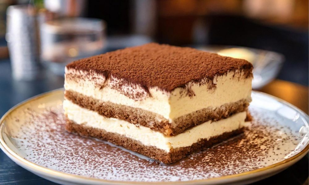 Quadratisches Stück Tiramisu mit Schichten aus cremiger Mascarpone und kaffeegesättigten Biskuits, oben dick mit Kakaopulver bestäubt und auf einem Teller in einem verschwommenen Café-Hintergrund serviert.