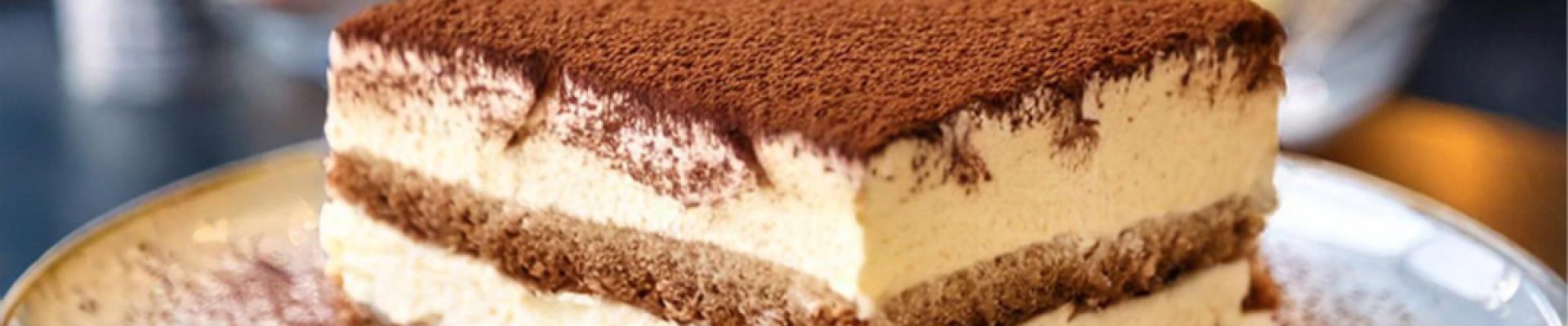 Quadratisches Stück Tiramisu mit Schichten aus cremiger Mascarpone und kaffeegesättigten Biskuits, oben dick mit Kakaopulver bestäubt und auf einem Teller in einem verschwommenen Café-Hintergrund serviert.
