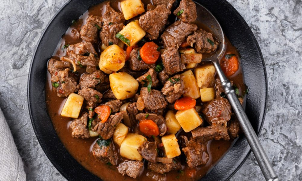 Schwarze Schüssel mit herzhaftem Rindergulasch: zarte Fleischwürfel, Kartoffel- und Möhrenstücke in brauner Sauce, Löffel im Rand und kleine Schale mit Sauerrahm im Hintergrund.