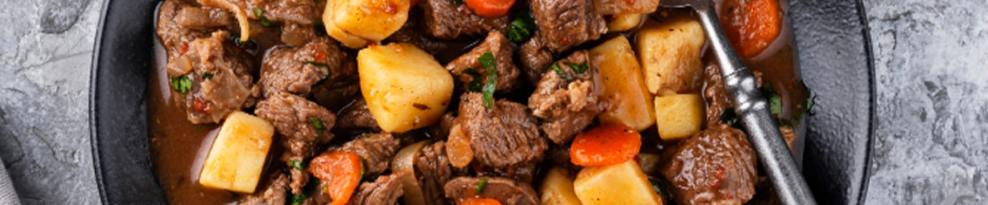 Schwarze Schüssel mit herzhaftem Rindergulasch: zarte Fleischwürfel, Kartoffel- und Möhrenstücke in brauner Sauce, Löffel im Rand und kleine Schale mit Sauerrahm im Hintergrund.