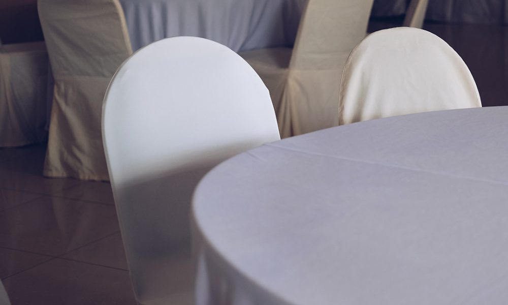 Leerer Bankettsaal mit mehreren runden, weiß gedeckten Tischen und Stühlen, die in cremefarbenen Hussen bezogen sind.