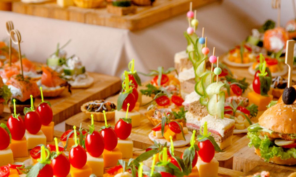 Vielseitiges Buffet mit farbenfrohen Canapés auf Holzplatten: Käsewürfel mit Kirschtomaten und Kräutern auf Spießen, Mini‑Sandwiches mit Gurkenspiralen, Tartes und ein kleines Burgerbrötchen im Hintergrund.