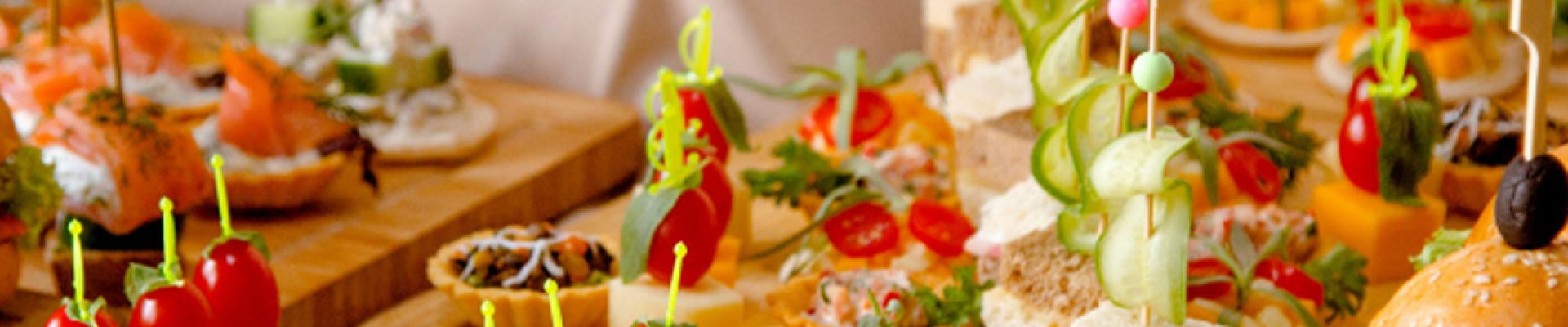 Vielseitiges Buffet mit farbenfrohen Canapés auf Holzplatten: Käsewürfel mit Kirschtomaten und Kräutern auf Spießen, Mini‑Sandwiches mit Gurkenspiralen, Tartes und ein kleines Burgerbrötchen im Hintergrund.