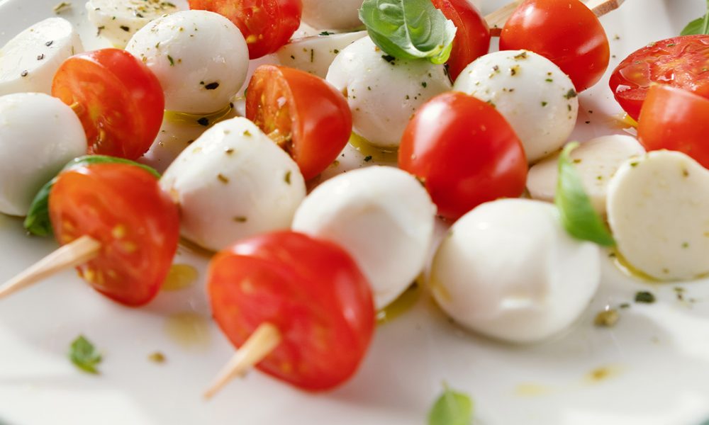 Spieße aus Kirschtomaten und Mozzarellakugeln auf einem weißen Teller, garniert mit Basilikum, Olivenöl und Kräutern.