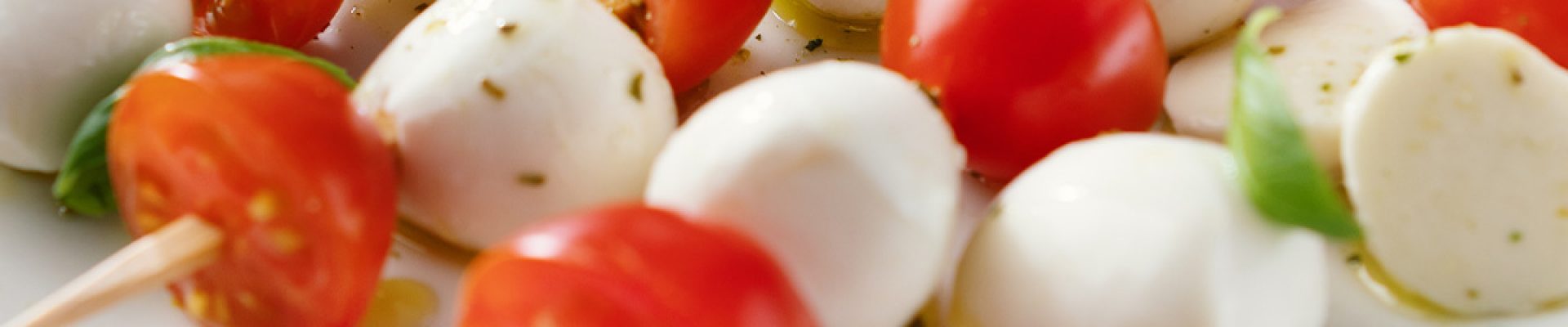 Spieße aus Kirschtomaten und Mozzarellakugeln auf einem weißen Teller, garniert mit Basilikum, Olivenöl und Kräutern.
