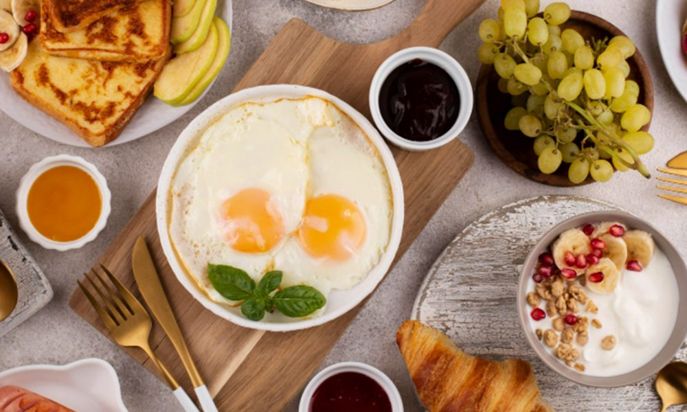 Vogelperspektive auf ein reichhaltiges Frühstück mit einer Schale Spiegeleier auf einem Holzbrett, Croissant, French Toast mit Apfelscheiben, Trauben, Joghurt mit Bananen, Granola, Würstchen, Butter sowie kleinen Schälchen mit Marmelade und Honig.