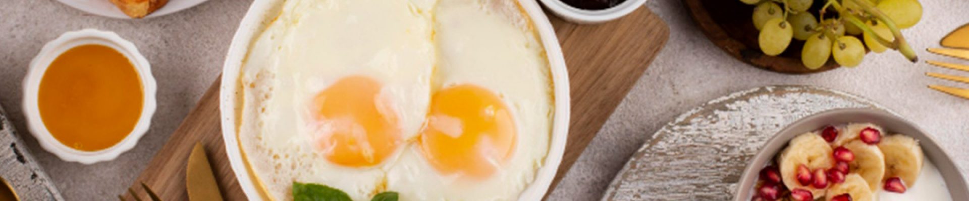 Vogelperspektive auf ein reichhaltiges Frühstück mit einer Schale Spiegeleier auf einem Holzbrett, Croissant, French Toast mit Apfelscheiben, Trauben, Joghurt mit Bananen, Granola, Würstchen, Butter sowie kleinen Schälchen mit Marmelade und Honig.