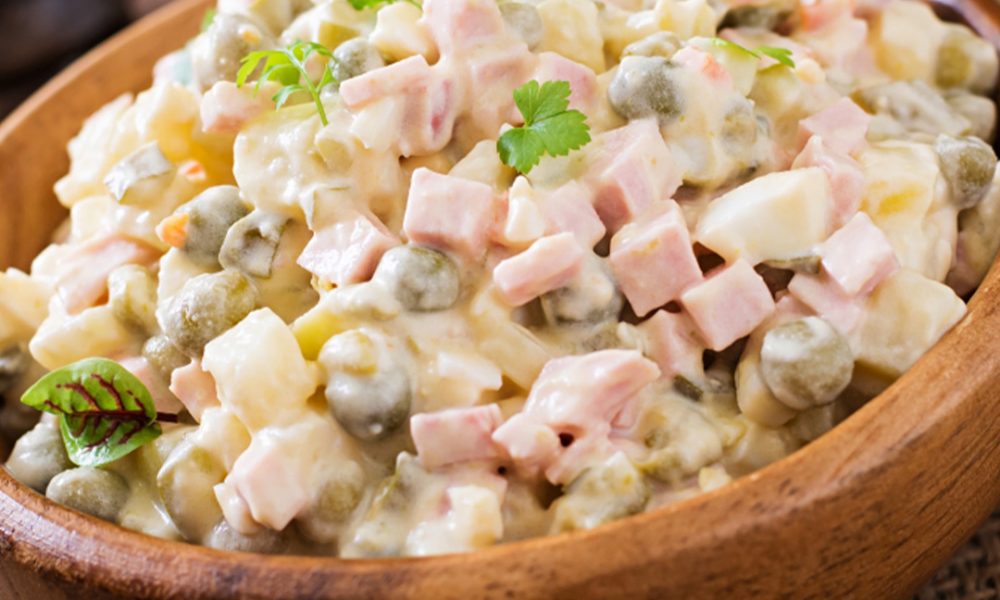 Holzschale mit russischem Salat (Olivier) aus gewürfeltem Schinken, Kartoffeln, Erbsen und cremiger Mayonnaise, garniert mit Petersilienblättchen.