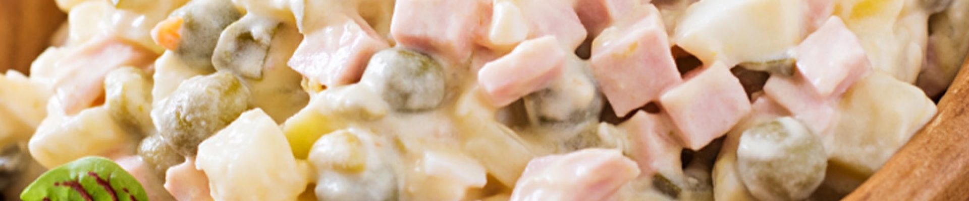 Holzschale mit russischem Salat (Olivier) aus gewürfeltem Schinken, Kartoffeln, Erbsen und cremiger Mayonnaise, garniert mit Petersilienblättchen.