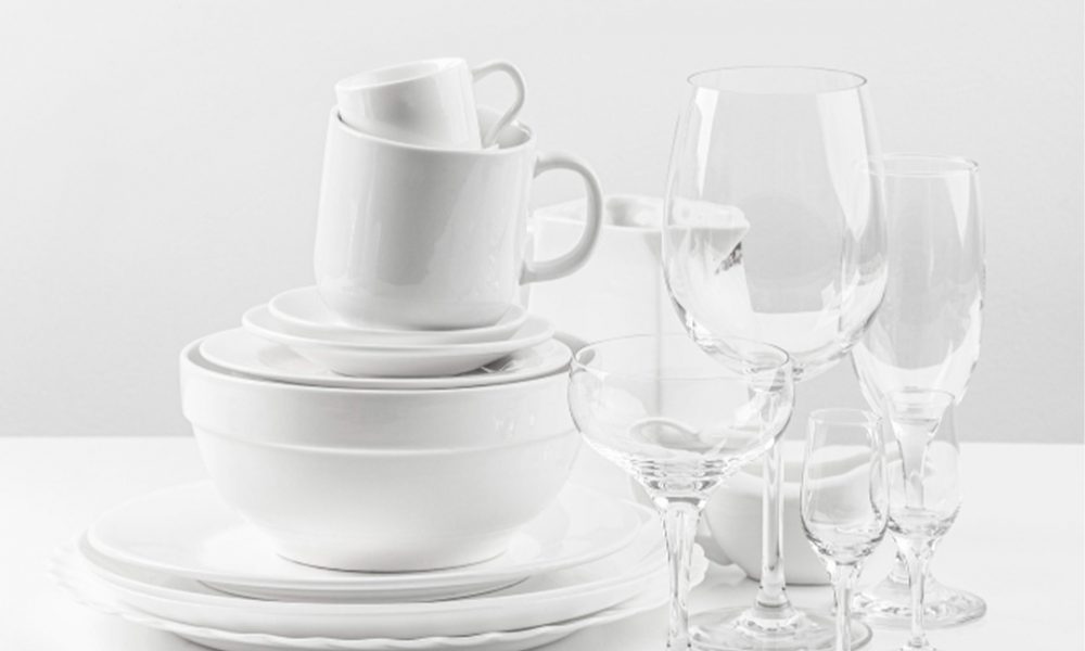 Gestapelte weiße Teller, Schüsseln und ineinander gesteckte Tassen links, daneben mehrere klare Weingläser und Sektgläser auf einem weißen, minimalistischen Hintergrund.
