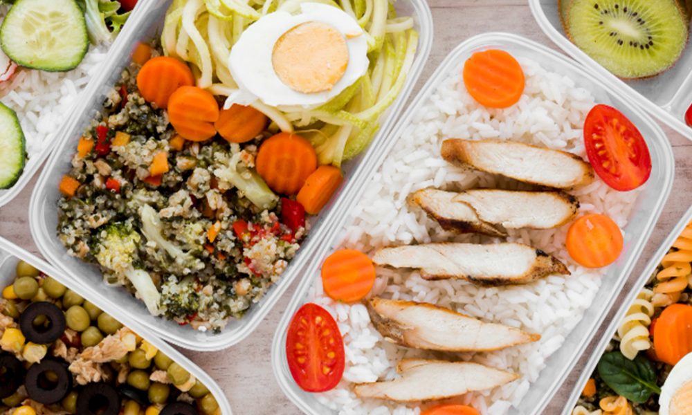 Mehrere Meal‑Prep‑Boxen mit gesunden Zutaten: eine mit Weißreis, in Streifen geschnittener gegrillter Hähnchenbrust, Karottenscheiben und Cherrytomaten; andere mit spiralisierten Zucchini und einem halben gekochten Ei, Quinoa‑Gemüse‑Mischung, Salaten, Oliven, Mais und Kiwischeiben.