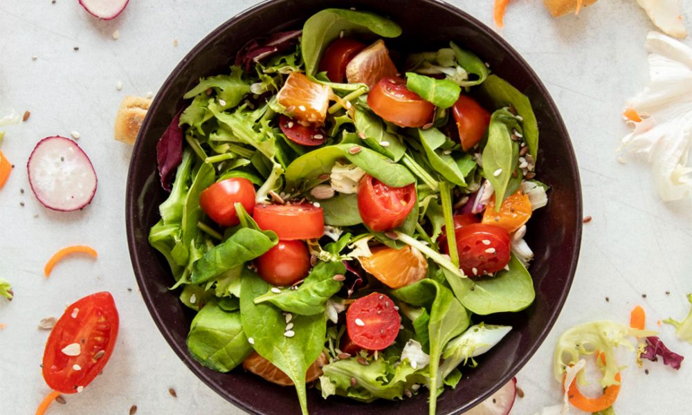 Schwarze Schüssel mit bunter Salatmischung aus Babyspinat und Blattsalaten, halbierten Kirschtomaten, Mandarinenstücken, Radieschenscheiben, Karottenröllchen, Croutons und Samen, auf hellem Untergrund mit verstreuten Salatbestandteilen.