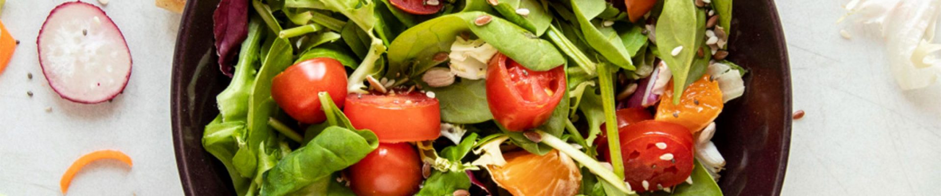 Schwarze Schüssel mit bunter Salatmischung aus Babyspinat und Blattsalaten, halbierten Kirschtomaten, Mandarinenstücken, Radieschenscheiben, Karottenröllchen, Croutons und Samen, auf hellem Untergrund mit verstreuten Salatbestandteilen.