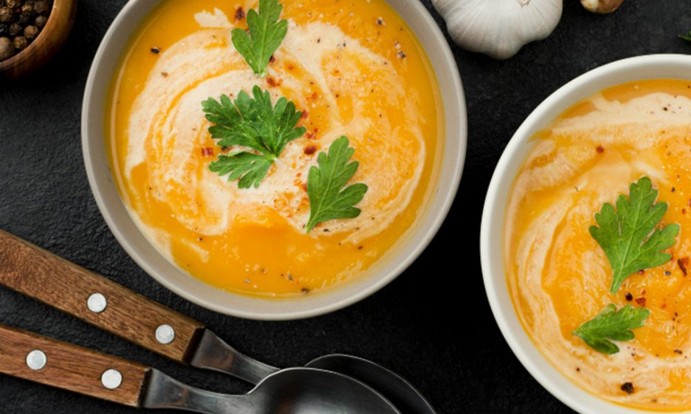 Zwei Schalen cremiger orangefarbener Suppe mit Sahneswirbeln und Petersilienblättern garniert, daneben zwei Metalllöffel mit Holzgriffen, Knoblauchzehen, Pfefferkörner und ein Leinentuch auf dunklem Schieferuntergrund.