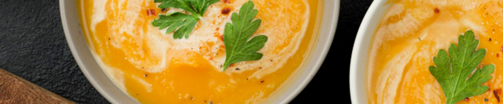 Zwei Schalen cremiger orangefarbener Suppe mit Sahneswirbeln und Petersilienblättern garniert, daneben zwei Metalllöffel mit Holzgriffen, Knoblauchzehen, Pfefferkörner und ein Leinentuch auf dunklem Schieferuntergrund.