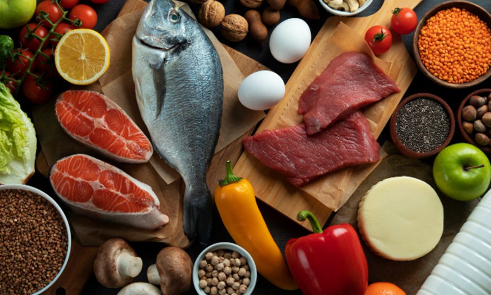 Verschiedene proteinreiche und frische Lebensmittel auf dunklem Holz angeordnet: ganzer Fisch und Lachssteaks, rohe Rindfleischfilets, Eier, Nüsse und Samen, Hülsenfrüchte, Pilze, Tomaten, Paprika, Äpfel, Zitrone, Käse und Milch.