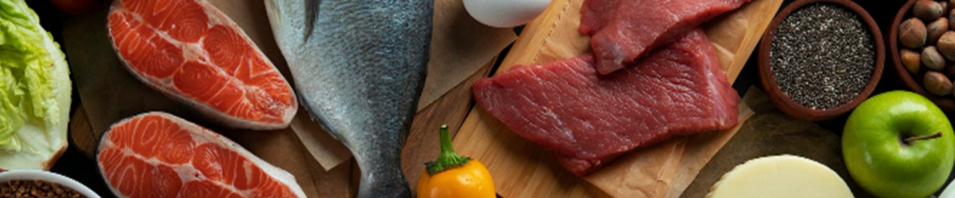 Verschiedene proteinreiche und frische Lebensmittel auf dunklem Holz angeordnet: ganzer Fisch und Lachssteaks, rohe Rindfleischfilets, Eier, Nüsse und Samen, Hülsenfrüchte, Pilze, Tomaten, Paprika, Äpfel, Zitrone, Käse und Milch.